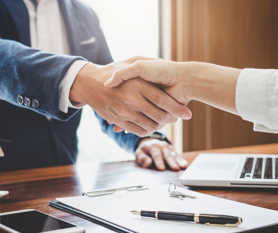 Two people shake hands over a paperwork filled desk as they finish a conversation focused on personal insurance guidance.